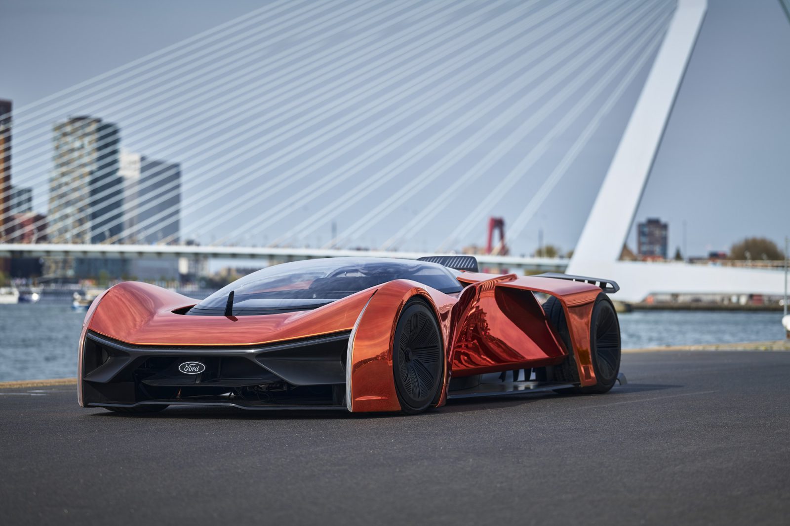 Een strakke, futuristische oranje Fordzilla P1-conceptauto staat geparkeerd op een weg aan het water, met een moderne brug en stadsgebouwen op de achtergrond.