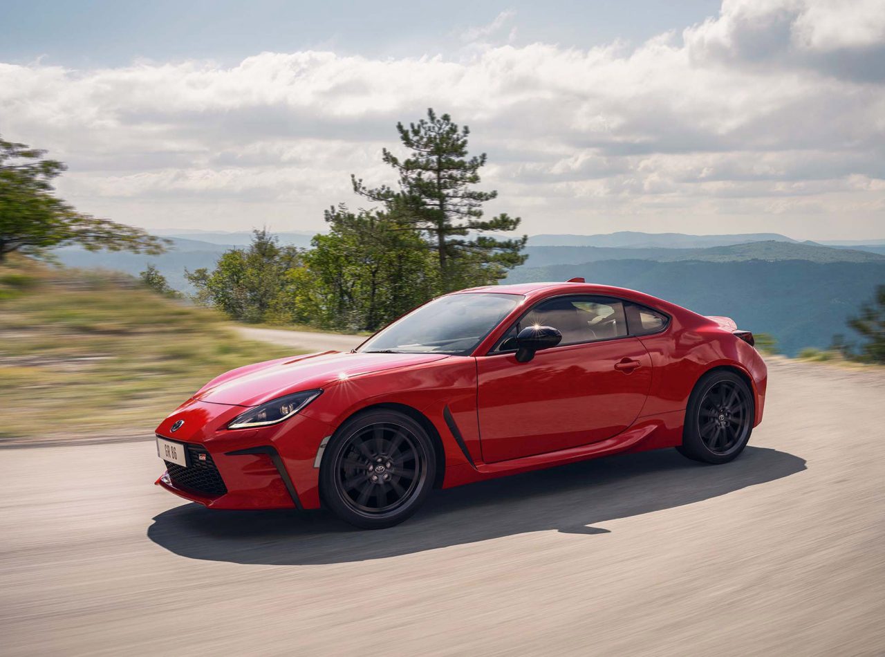 Een Toyota GR86, met zijn strakke rode ontwerp, snelt over een bergachtige weg omgeven door groen en bomen onder een bewolkte hemel.