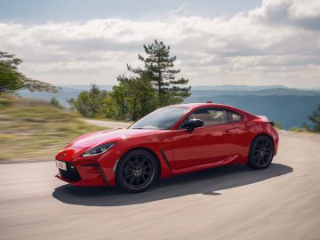 Een Toyota GR86, met zijn strakke rode ontwerp, snelt over een bergachtige weg omgeven door groen en bomen onder een bewolkte hemel.