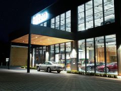 Een moderne autodealer bij nacht, met een glazen gevel waarbinnen verschillende auto's te zien zijn, die lijkt op een autohotel. Onder de verlichte ingang staat een klassieke witte auto geparkeerd.