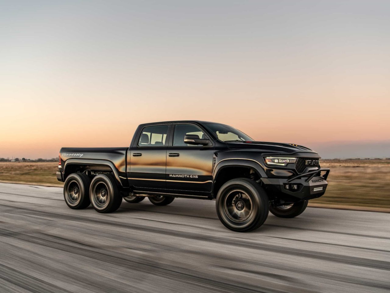 Een zwarte Mammoth 1000 6x6 TRX pick-uptruck rijdt op een verharde weg met een vlak landschap en een zonsonderganghemel op de achtergrond.