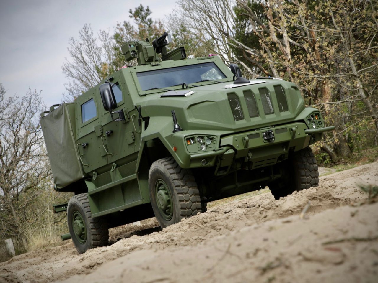 Een olijfgroen gepantserd militair voertuig van Manticore staat geparkeerd op zanderig terrein, omgeven door bomen.