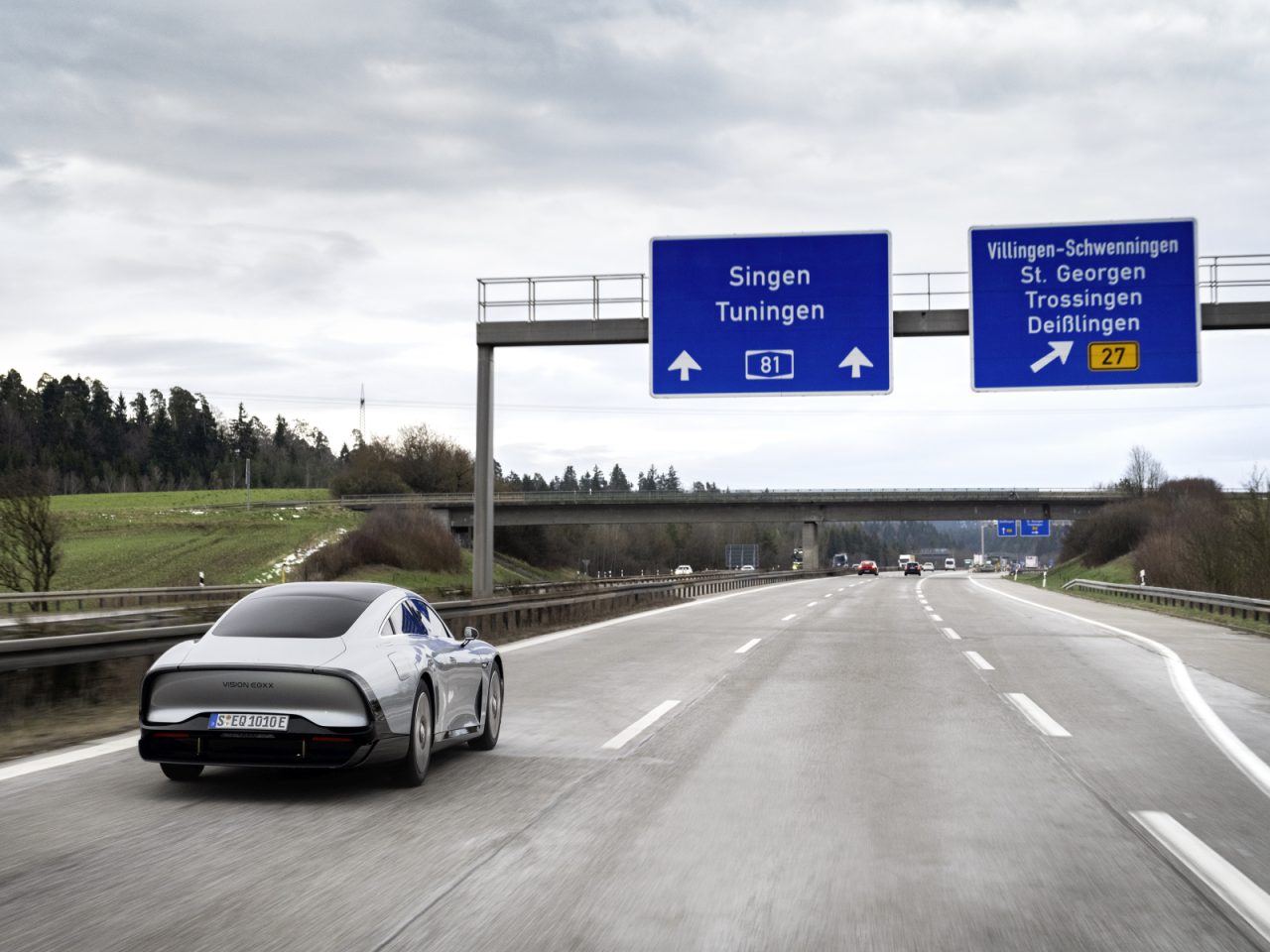 Een futuristische Mercedes EQXX rijdt op een snelweg onder bewolkte hemel. Bovengrondse borden geven de richting Singen en Villingen-Schwenningen aan.