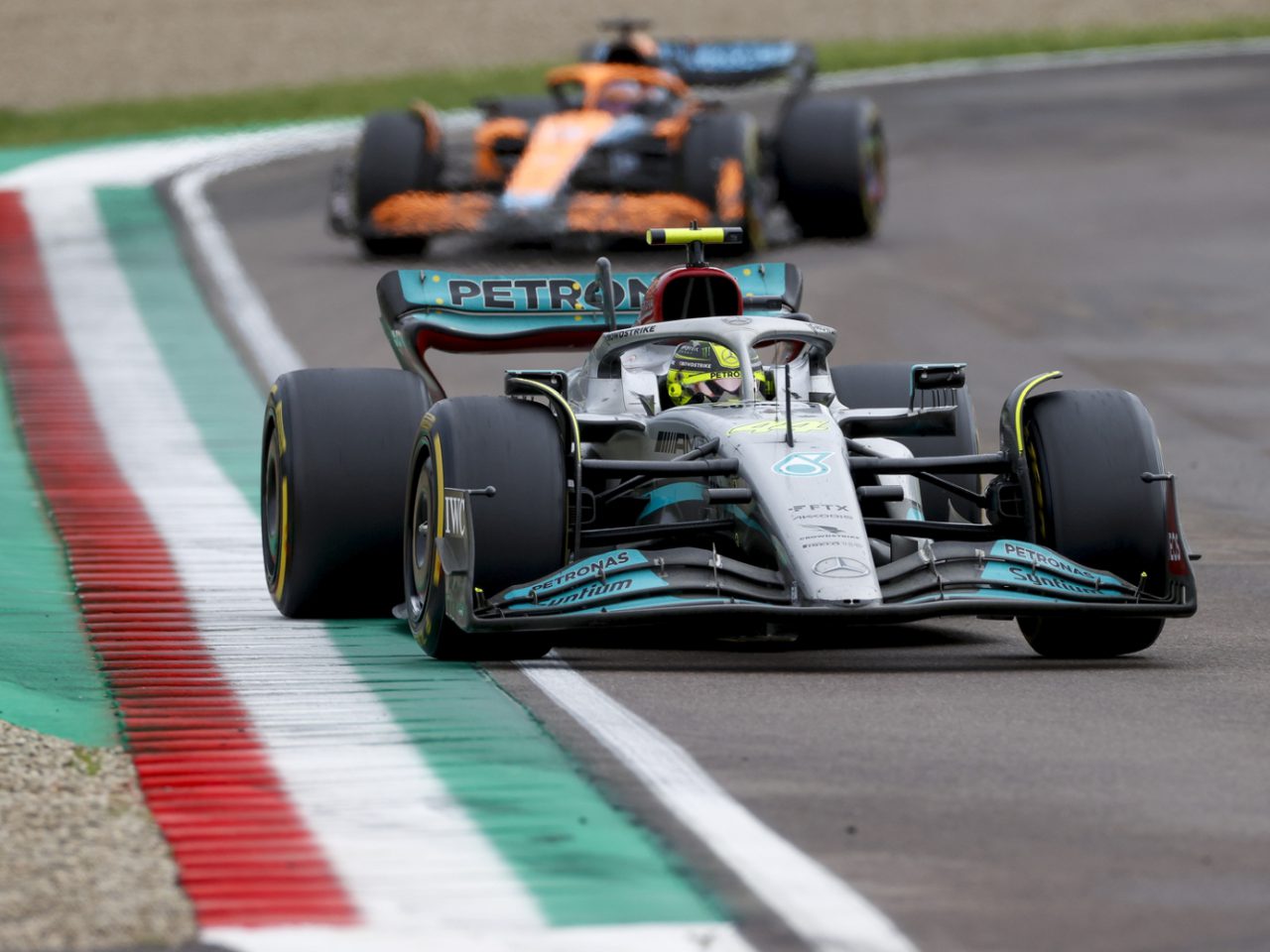 Een raceauto met het Petronas-logo, bestuurd door George Russell, leidt op een circuit, op de voet gevolgd door een andere auto. De banden van de leidende auto staan gedeeltelijk op een rode, witte en groene stoeprand.