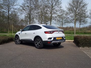 Een strakke witte Renault Arkana-auto staat geparkeerd op een buitenterrein, omgeven door bladerloze bomen en groen. Het uitzicht geeft op sierlijke wijze de achterkant en bestuurderszijde van het stijlvolle voertuig weer.