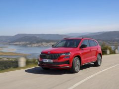 Een rode Skoda Karoq SUV rijdt op een schilderachtige weg met bergen en een meer op de achtergrond onder een helderblauwe hemel en biedt een onberispelijke achtergrond voor deze recensie.