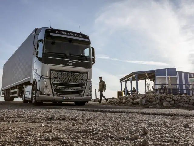 Een grote witte Volvo FH-truck staat geparkeerd op een grindweg bij een klein gebouw, terwijl er vlakbij een man loopt. Op de voorkant van de vrachtwagen staat het woord