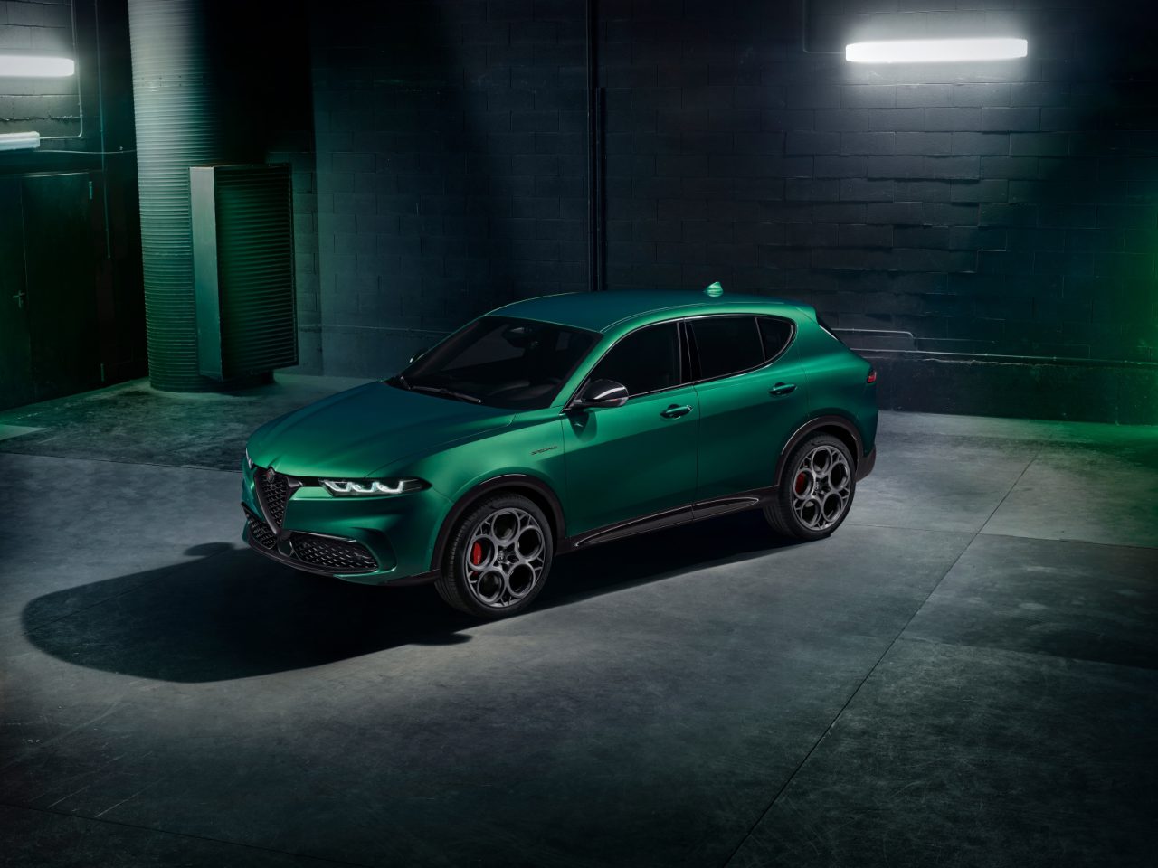 Een groene Alfa Romeo Tonale SUV staat binnen geparkeerd in een slecht verlichte kamer met betonnen vloeren en bakstenen muren. Twee fluorescentielampen erboven zorgen voor een subtiele verlichting van het gestroomlijnde voertuig.