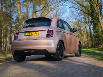 Een roze Fiat 500e met zwarte wielen staat geparkeerd op een geplaveid pad vlakbij een bosrijke omgeving. Op het kenteken staat "L-583-PL.