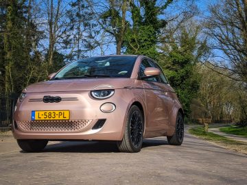 Een lichtbruine Fiat 500e compacte auto met kenteken "L-583-PL" staat geparkeerd op een verharde weg omgeven door bomen.