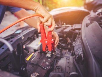 Een persoon gebruikt rode startkabels om de accu van een accu leeg auto te starten. De motorkap van de auto is open en de zon schijnt op de achtergrond.