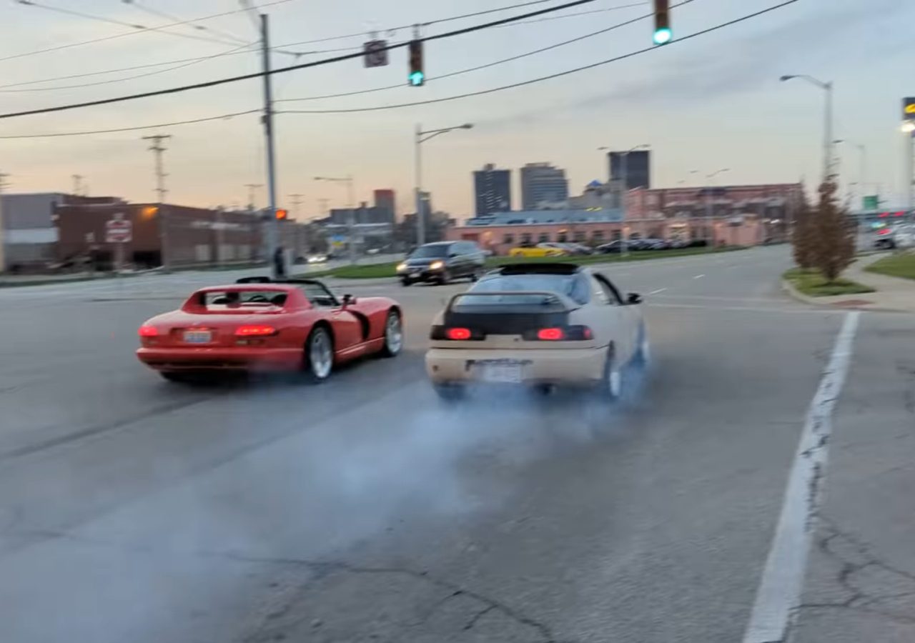 Twee sportwagens, een rode Dodge Viper en een witte coupé, accelereren vanaf een kruispunt en laten een rookspoor achter. Op de achtergrond zijn stedelijke gebouwen en verkeerslichten zichtbaar.