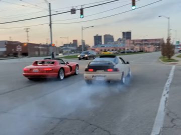 Twee sportwagens, een rode Dodge Viper en een witte coupé, accelereren vanaf een kruispunt en laten een rookspoor achter. Op de achtergrond zijn stedelijke gebouwen en verkeerslichten zichtbaar.