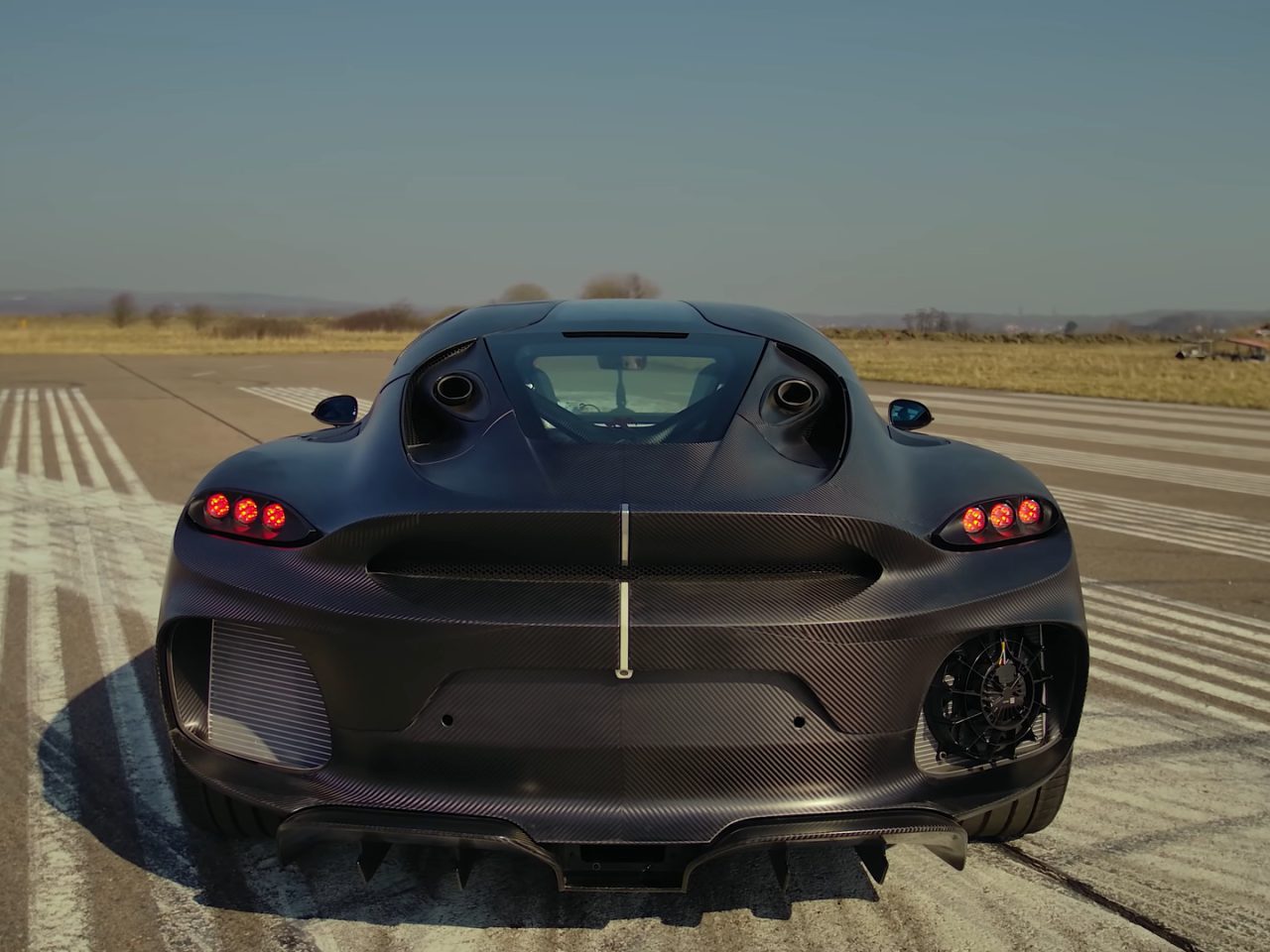 Een zwarte Koenigsegg-sportwagen staat geparkeerd op een lege landingsbaan met zichtbare strepen op de stoep. De camera legt het achteraanzicht vast en benadrukt de achterlichten, het strakke ontwerp en het dubbele uitlaatsysteem van de auto.