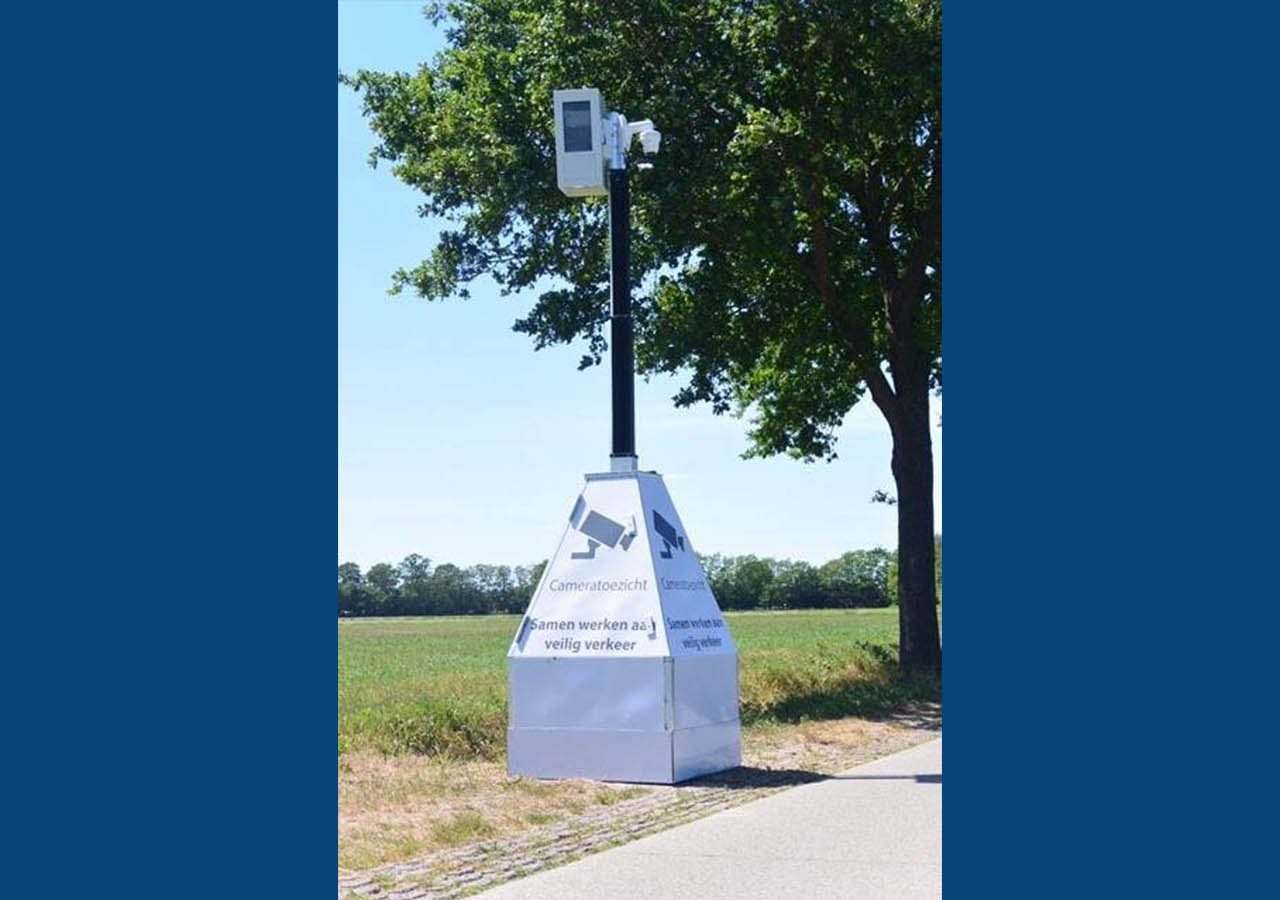 Aan de kant van een landelijke weg naast een boom staat een hoge flitspalenflitspaal, met een witte, doosachtige basis waarop tekst en symbolen worden weergegeven.