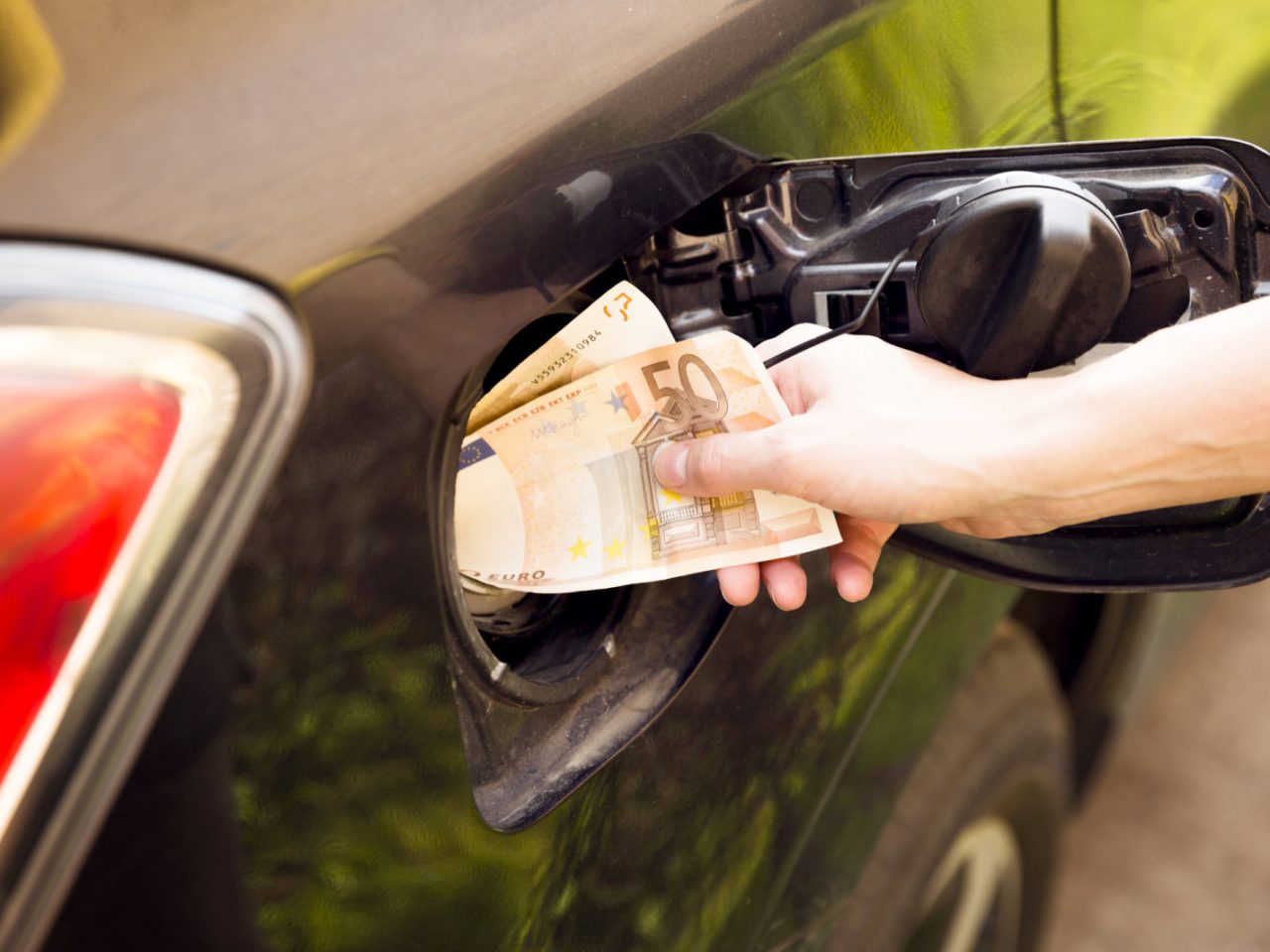 Een hand stopt eurobankbiljetten in de brandstoftank van een auto, met de tankdop open, wat de torenhoge benzineprijs symboliseert.