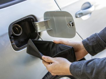 Persoon die een open, lege portemonnee vasthoudt bij een open deur van de benzinetank van een auto, die de impact van de stijgende benzineprijs op het dagelijks leven weerspiegelt.