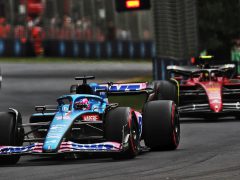 Op het circuit van Emilia Romagna racen twee Formule 1-auto's. De auto op de voorgrond is blauw en de auto erachter is rood. Beide auto's navigeren door een bocht, waarbij bandenbarrières en toeschouwers op de achtergrond zichtbaar zijn.