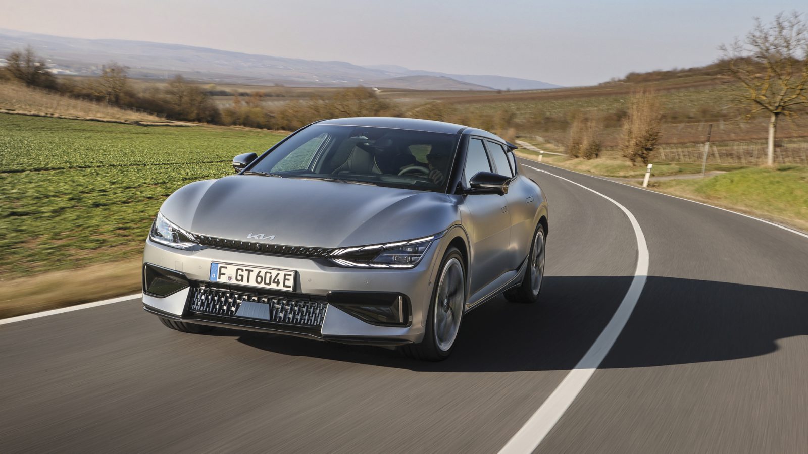 Een zilveren Kia EV6 GT rijdt over een bochtige weg door een landelijk landschap met groene velden en kale bomen onder een heldere hemel.