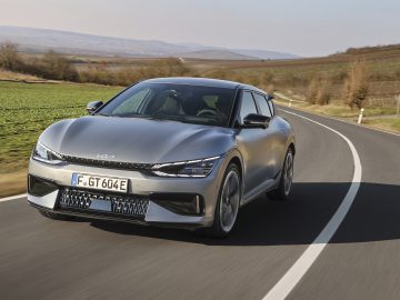 Een zilveren Kia EV6 GT rijdt over een bochtige weg door een landelijk landschap met groene velden en kale bomen onder een heldere hemel.