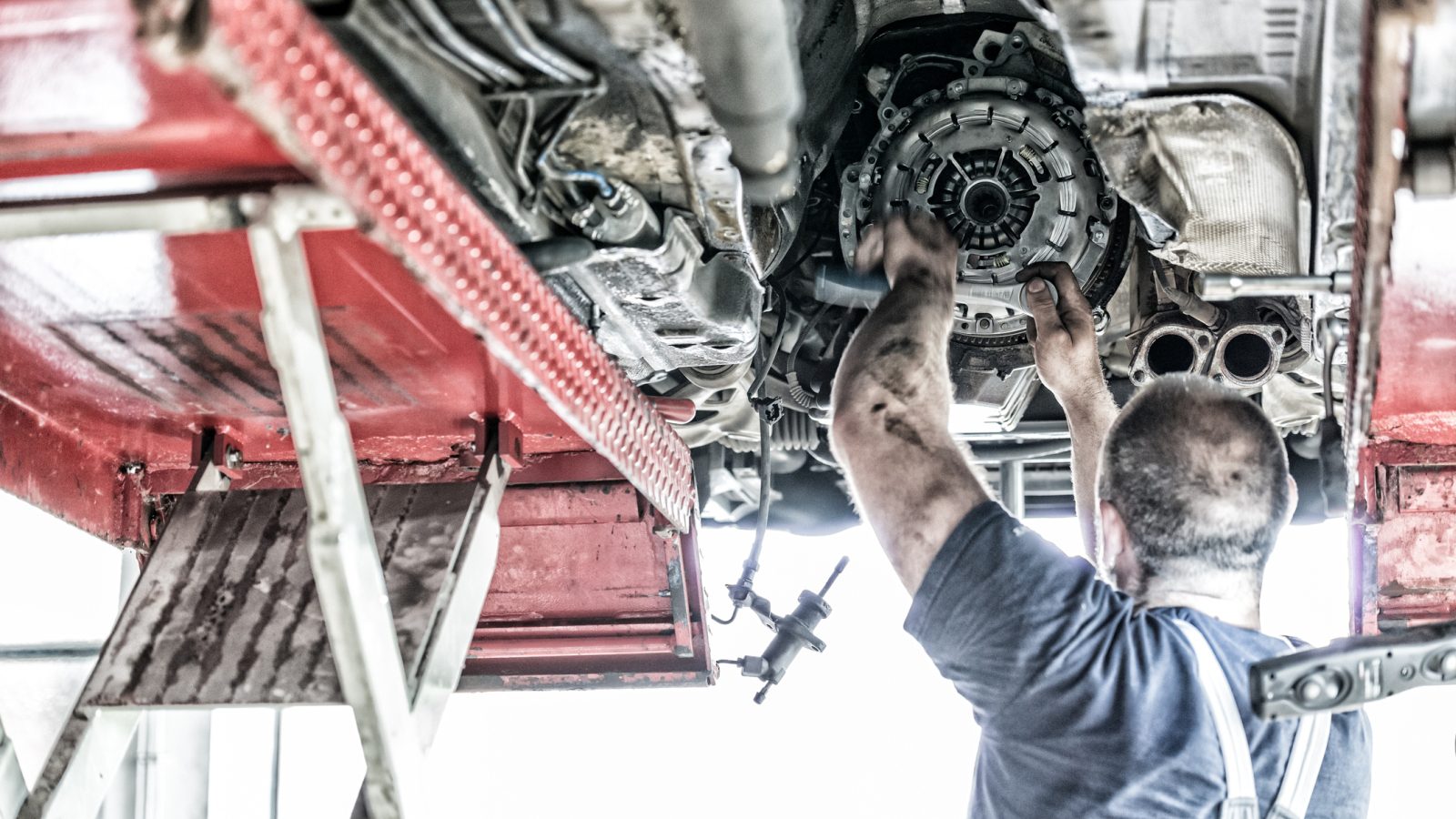 Een monteur werkt aan de onderkant van een voertuig dat op een autolift wordt gehesen, waarbij hij onderdelen in de buurt van de motor aanpast om een probleem met de slippende koppeling op te lossen.