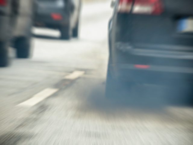 Een close-upfoto van een zwarte auto die uitlaatgassen uitstoot, wat wijst op de noodzaak van een roetfilter, terwijl hij op een weg rijdt met andere voertuigen op de achtergrond.