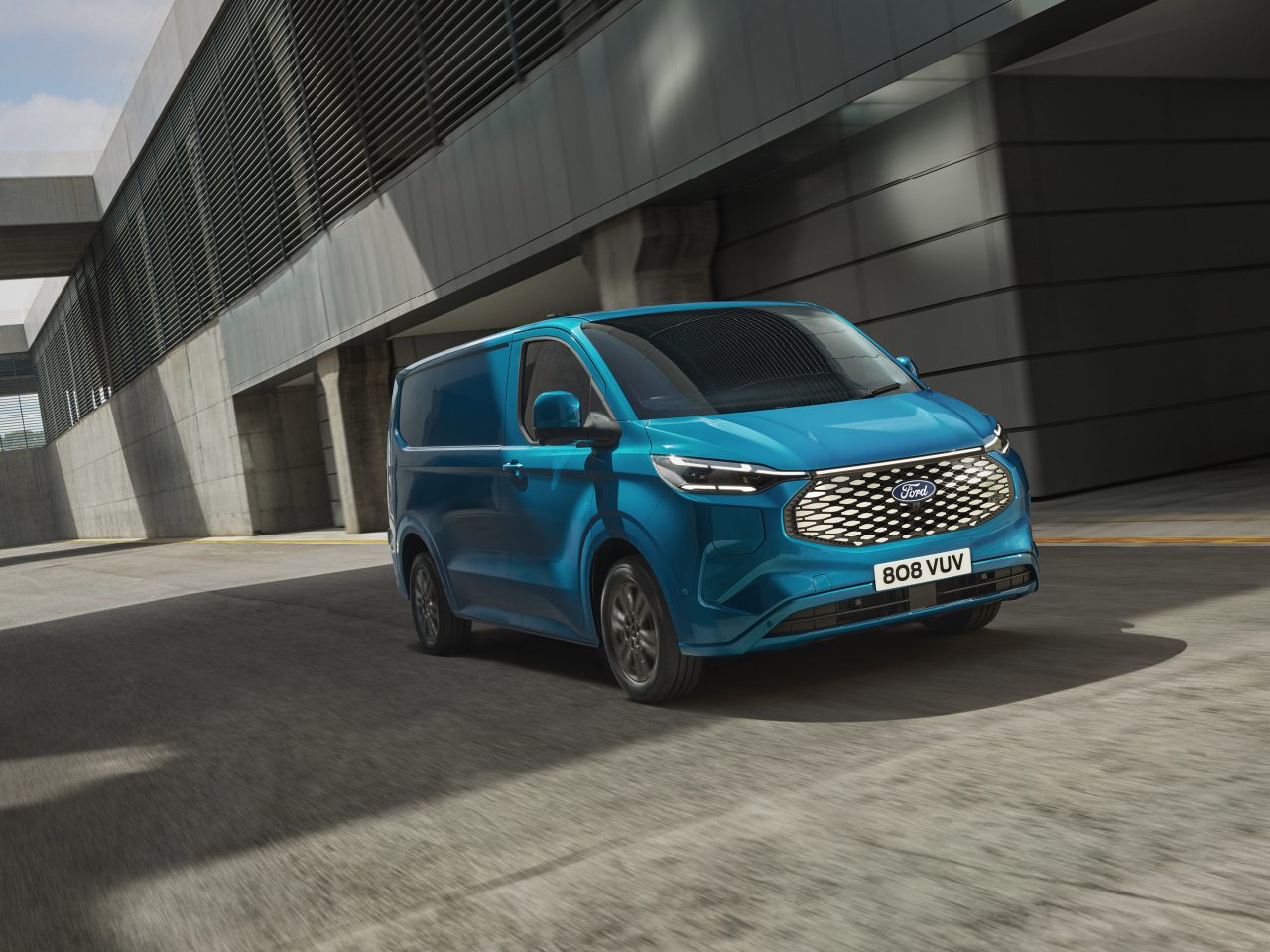Een blauwe Ford Transit rijdt door een modern stedelijk industriegebied. Het busje rijdt over een lege weg met aan weerszijden hoge betonnen gebouwen.