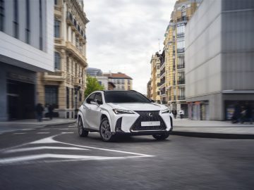 Een strakke witte Lexus UX glijdt door een stadsstraat die wordt begrensd door zowel historische als moderne gebouwen, met enkele voetgangers op de achtergrond.