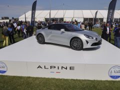 Een zilveren Alpine-sportwagen wordt prominent tentoongesteld op een verhoogd platform tijdens een buitenevenement, met een menigte en witte tenten op de achtergrond, waardoor ieders aandacht wordt getrokken.