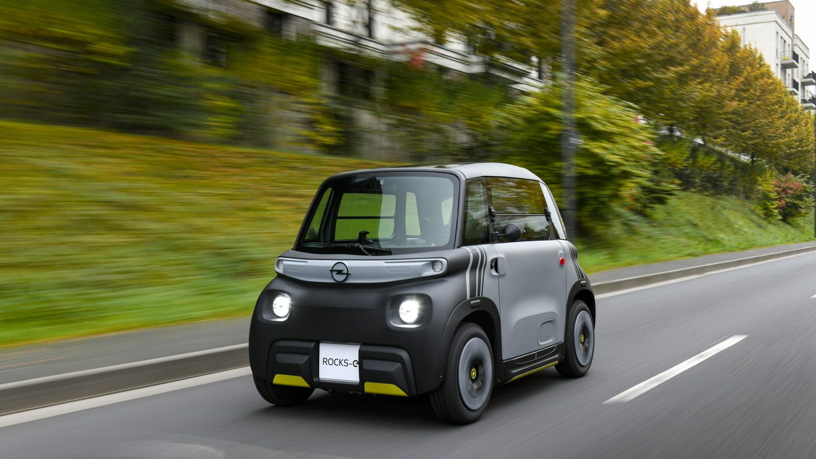 Een compacte tweedeurs Opel Rocks-e rijdt over een weg, met groen en gebouwen op de achtergrond.