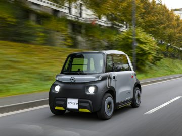 Een compacte tweedeurs Opel Rocks-e rijdt over een weg, met groen en gebouwen op de achtergrond.