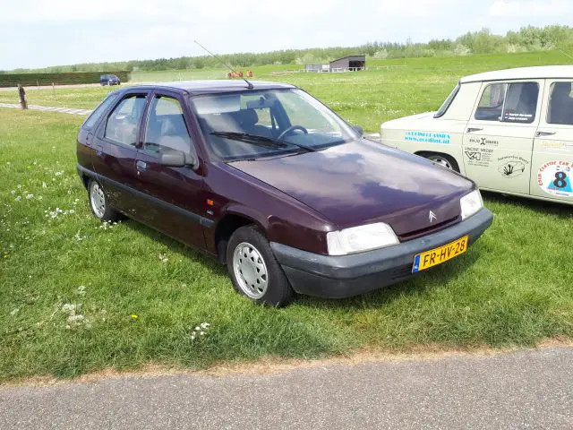 Een bordeauxrode Citroën ZX-hatchback, die de charme van Elk Merk Waardig tentoonspreidt, staat geparkeerd op grasveld naast een andere oldtimer met een blauw-oranje sticker. Op de achtergrond zijn enkele structuren en bomen zichtbaar.