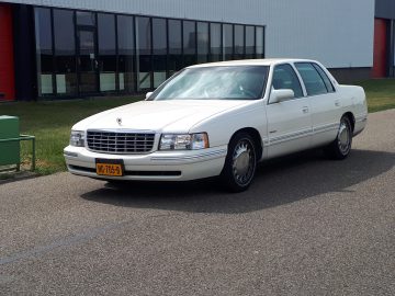 Een witte luxe sedan, die de eland merkwaardig standaard uitstraalt, staat geparkeerd op een weg naast een groen en wit industrieel gebouw. Het kenteken van de auto is zichtbaar en toont "HB-70S-8.