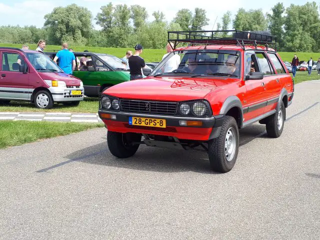 Een rode terreinwagen met kenteken 28-GPS-8 staat tijdens een auto-evenement van Elk Merk Waardig op een weg geparkeerd, omringd door mensen en andere geparkeerde auto's. Op de achtergrond zijn bomen en groen zichtbaar.