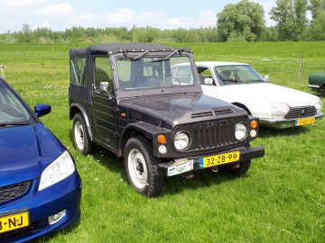 Een zwarte Suzuki-terreinwagen met een softtop, symbolisch voor de ruige geest van Elk Merk Waardig, staat geparkeerd op het gras naast een wit voertuig en een blauw voertuig. Op het kenteken aan de voorkant staat "32-ZB-99".