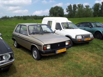 Drie geparkeerde auto's op een grasveld: een lichtbruine hatchback, een wit busje en een groene compacte auto, met bomen en een bewolkte lucht op de achtergrond. De scène belichaamt Elk Merk Waardig en legt rust en eenvoud midden in de natuur vast.