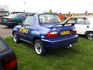 Een blauwe Suzuki X-90 met een achterspoiler en op maat gemaakte emblemen staat buiten op gras geparkeerd. Op de achtergrond zijn verschillende mensen te zien en er staat een ander voertuig vlakbij geparkeerd. Op het kenteken staat NV-LN-68. De scène straalt een Elk Merk Waardig-sfeer uit met zijn unieke auto-flair.