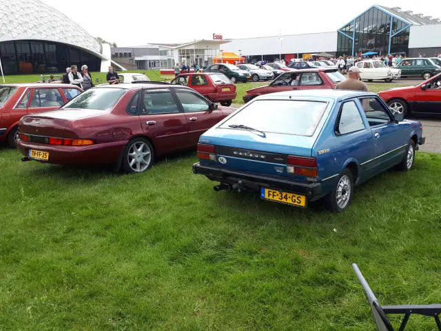 Twee oldtimers, een rode en een blauwe, staan geparkeerd op een grasveld tijdens de autoshow Elk Merk Waardig. Beide voertuigen hebben gele Europese kentekenplaten en op de achtergrond staat een groep mensen bij een gebouw.