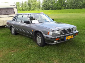 Een grijze sedan uit de jaren 80 geparkeerd op het gras naast een caravan, met een bordje 'Elk Merk Waardig' Te Koop op de voorruit. Nederlands kenteken SL-44-XX. Bomen en groen op de achtergrond.