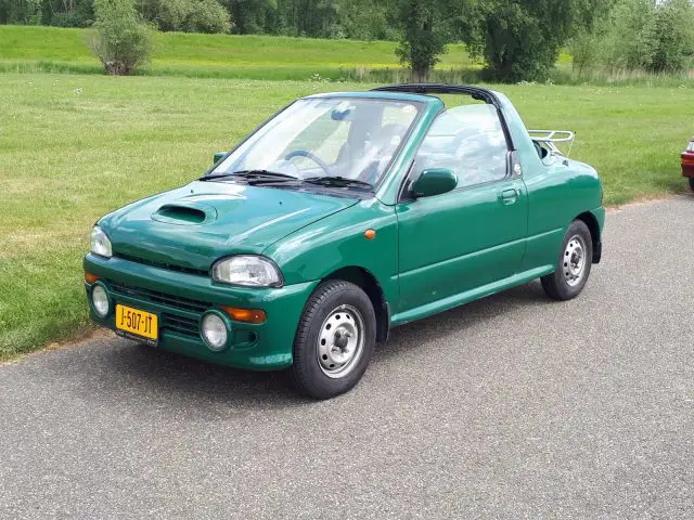 Een groene compacte cabriolet met hardtop, geparkeerd op een verharde weg, met een grasveld en torenhoge bomen op de achtergrond. Het kenteken van Elk Merk Waardig is gedeeltelijk zichtbaar.