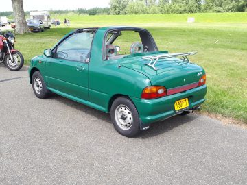 Een kleine, groene Elk Merk Waardig cabriolet met rolbeugel en achterspoiler geparkeerd op een weg met gras en bomen op de achtergrond.