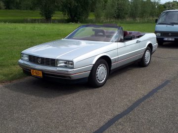 Een zilverkleurige cabriolet met geel kenteken staat geparkeerd op een verharde weg naast een groen grasveld met bomen op de achtergrond, wat de geest van Elk Merk Waardig belichaamt. Achter de auto staat een wit busje geparkeerd.