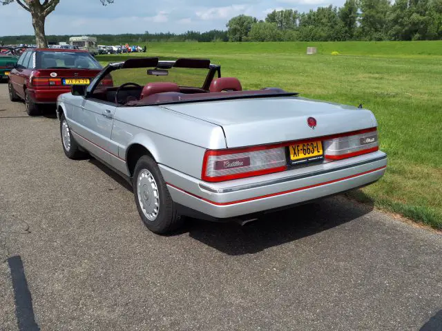 Een zilveren cabriolet Cadillac, die de kwaliteit van Elk Merk Waardig tentoonspreidt, staat buiten geparkeerd op een verhard terrein. De achtergrond bestaat uit groene velden en bomen. Het voertuig heeft Nederlandse kentekenplaten en een kastanjebruin interieur, met een andere auto gedeeltelijk zichtbaar in het frame.