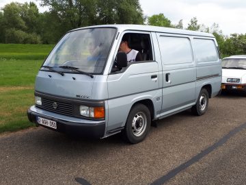 Een zilveren Nissan-busje met een man op de bestuurdersstoel, geparkeerd op een weg met gras en bomen op de achtergrond, beeldt een scène uit Elanden Merk Waardig af.