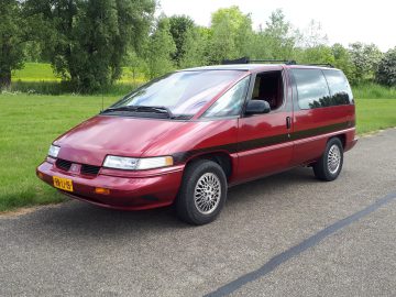 Een rode minibus, voorzien van een sticker van Elk Merk Waardig, staat geparkeerd op een verharde weg met groene bomen en gras op de achtergrond. De zijdeur staat open en aan de voorzijde is een gele kentekenplaat zichtbaar.