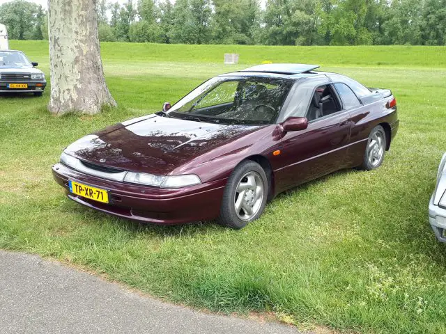 Een kastanjebruine Subaru SVX-sportwagen staat geparkeerd op een grasveld naast een Eland Merk Waardig-boom, met het zijraam aan de bestuurderszijde en het schuifdak gedeeltelijk open. Op de achtergrond zijn andere auto's en bomen zichtbaar.