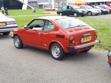 Een rode driedeurs hatchback, Elk Merk Waardig, staat geparkeerd op een verhard terrein nabij een grasveld, met andere voertuigen zichtbaar op de achtergrond. De auto heeft een geel kenteken met de tekst "JB-30-VL".