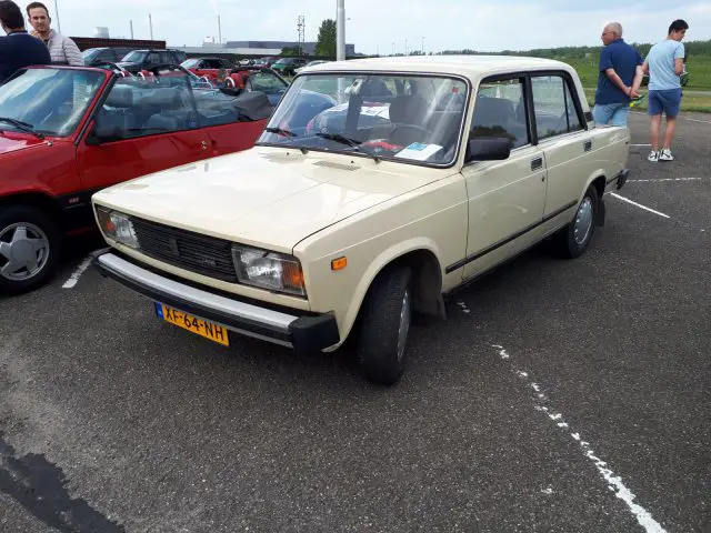 Op een parkeerplaats staat een beige klassieke auto met Nederlands kenteken, die de essentie van Elk Merk Waardig belichaamt. Er zijn mensen en andere auto's op de achtergrond.