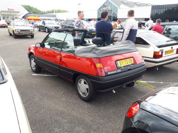 Een rode Renault 5 cabriolet, echt Elk Merk Waardig, staat tussen andere voertuigen geparkeerd op een buitenautoshow. Er staan een aantal mensen omheen, die een gesprek voeren.