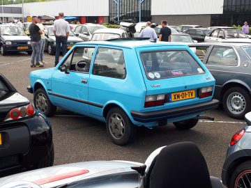 Een lichtblauwe hatchback met geel kenteken "XN-01-TV" staat tussen andere auto's geparkeerd op een buitenbijeenkomst en belichaamt de essentie van Elk Merk Waardig, met verschillende mensen die op de achtergrond staan te praten.